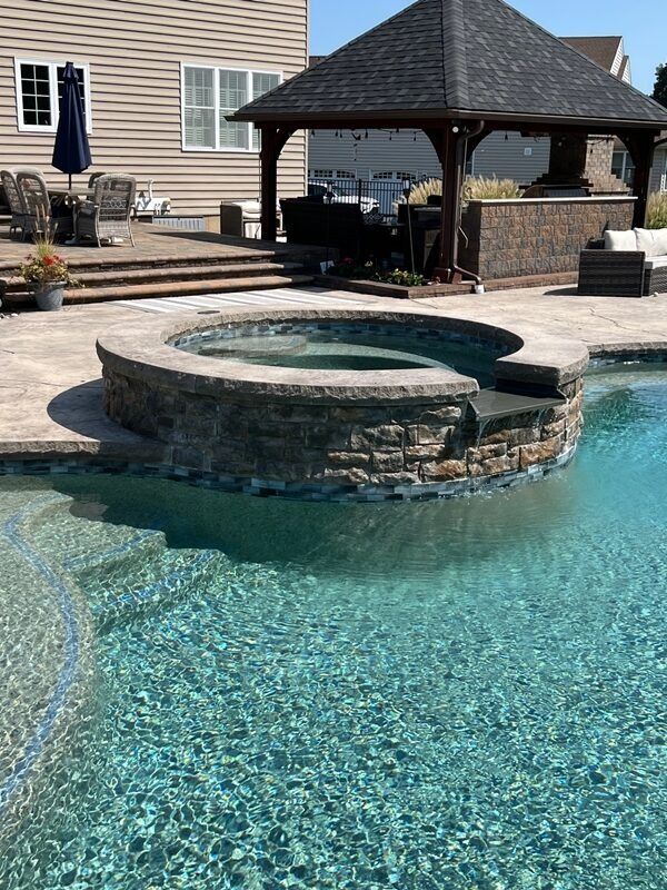 Pool with built-in hot tub, steps, and gazebo with an outdoor kitchen, on a sunny day.