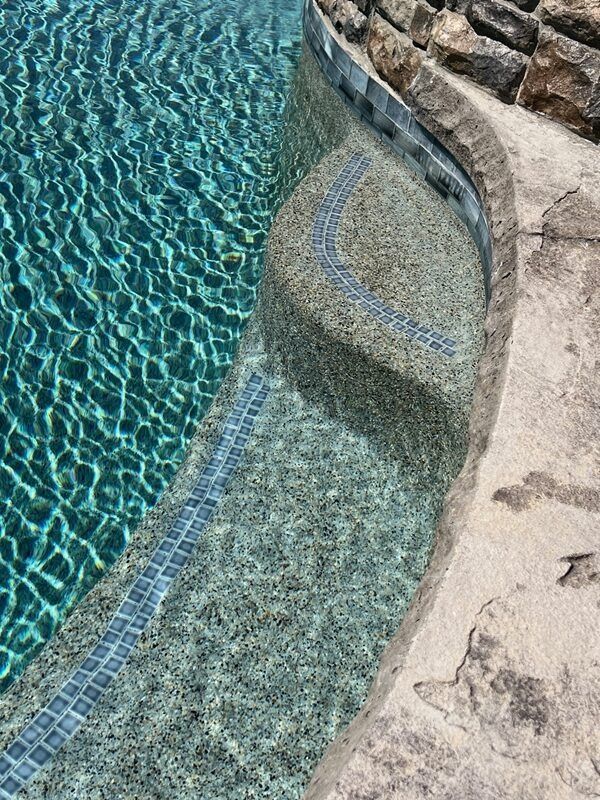 Close-up of a pool with steps. Turquoise water, pebble finish on steps, blue tile detail. Stone and concrete surround.