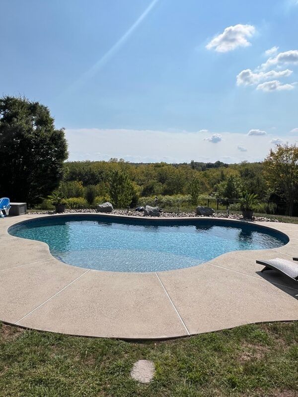 Pool with blue water surrounded by concrete patio, green grass, and trees under a blue sky.