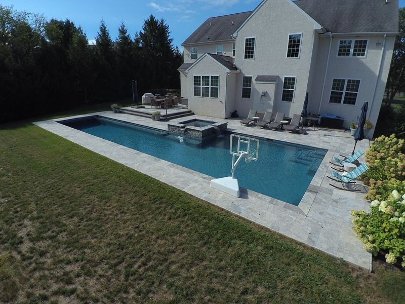 Backyard with rectangular pool, spa, and basketball hoop. House in background, blue sky.