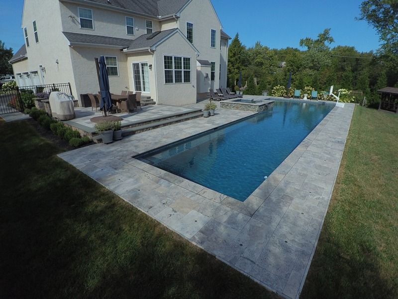 Backyard with rectangular pool and house. Gray stone patio, blue water, green grass, and trees in background.