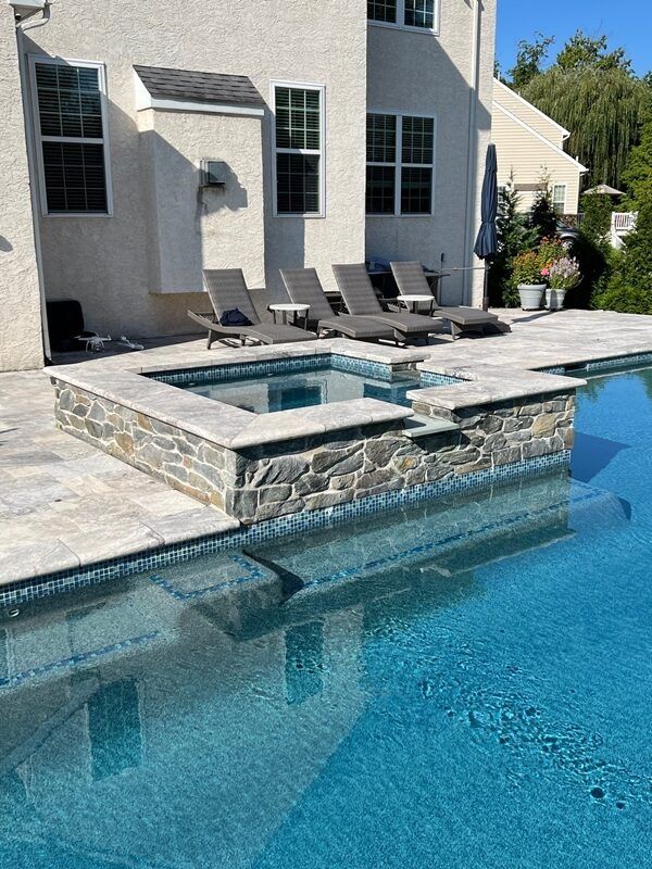 Swimming pool with a stone hot tub, lounge chairs, and a house in the background. Blue water and a sunny day.