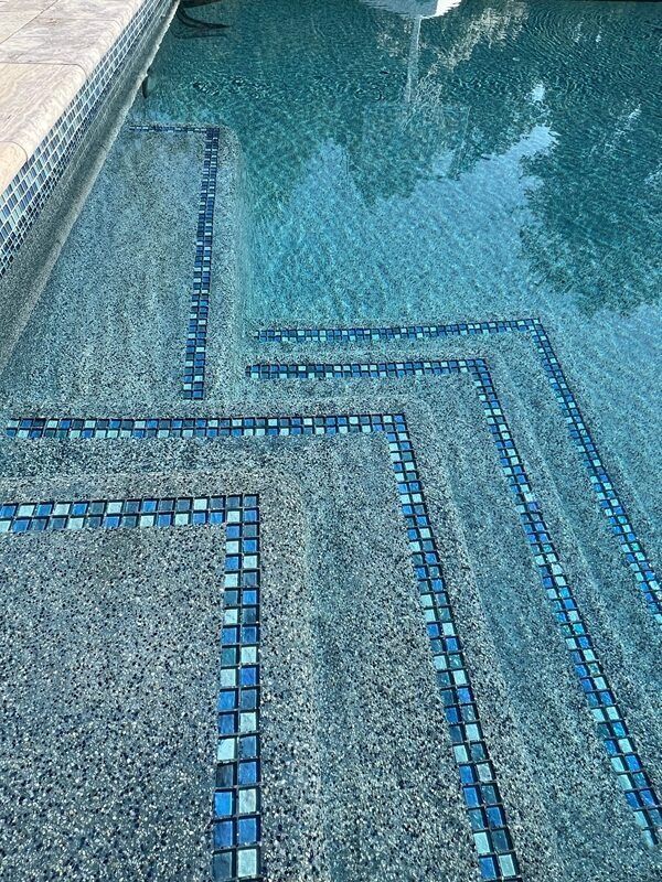 Pool with step design, blue and gray mosaic tiles, water.