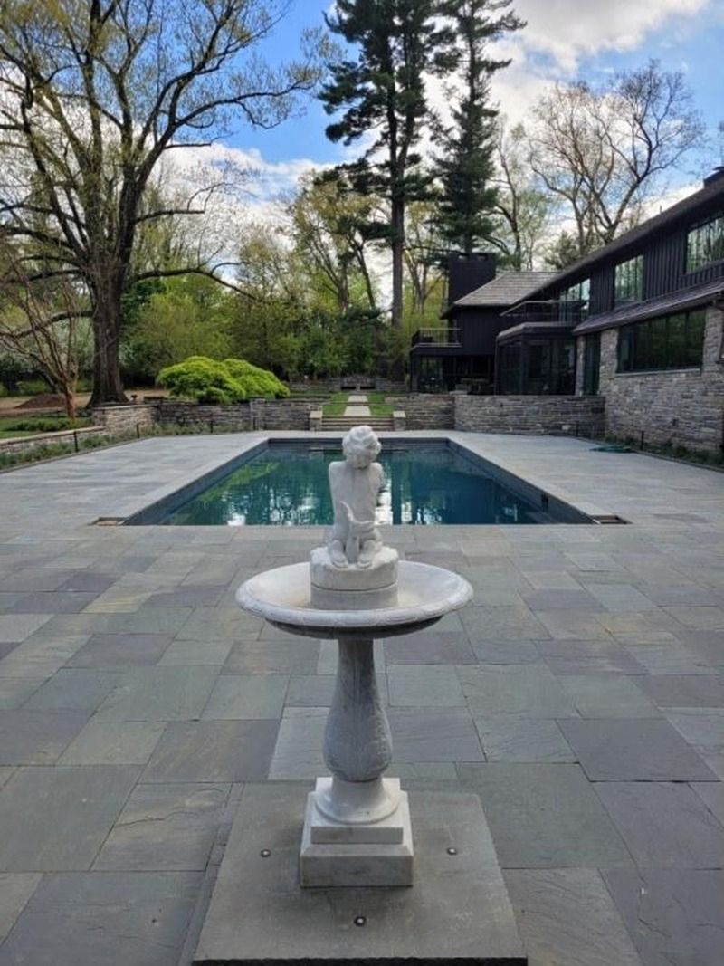 Marble fountain in front of a rectangular pool, dark house in background, gray stone patio, trees and blue sky.