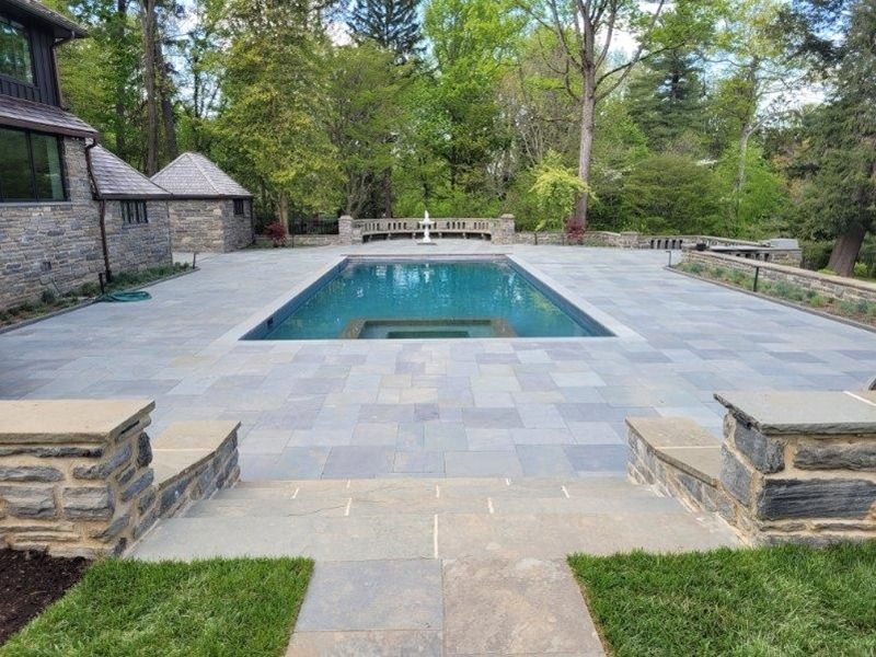 Stone patio with rectangular pool, seating areas, and a small fountain. Surrounded by green lawn and trees.