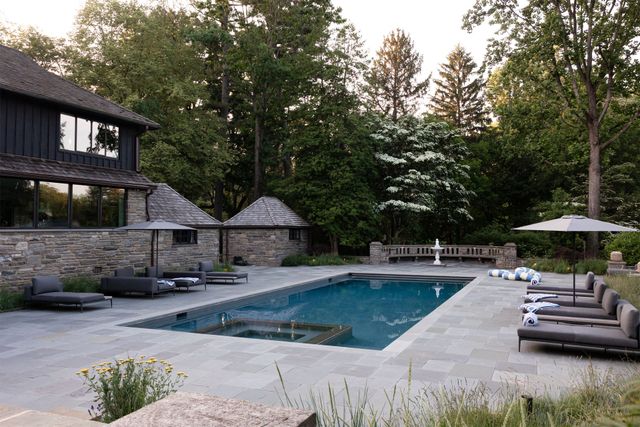 Modern backyard with a rectangular swimming pool, lounge chairs, and trees beside a stone house