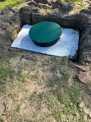 Green round septic tank cover in a dug-out pit on a white slab in a grassy yard