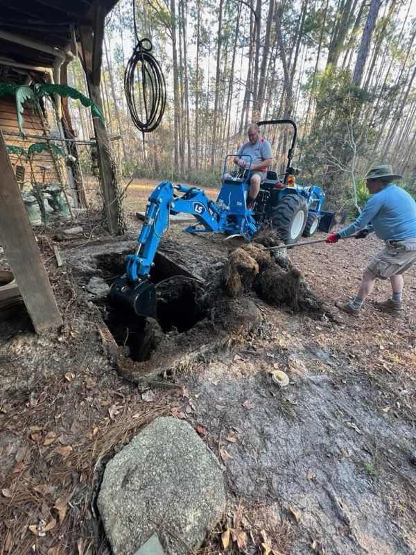 Blue backhoe digging a large hole in a wooded area with two workers nearby