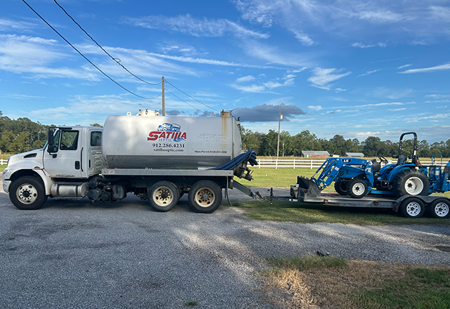Learn More About Why Clean My Tank? White tanker truck towing a blue tractor on a trailer parked outside on a cloudy day.