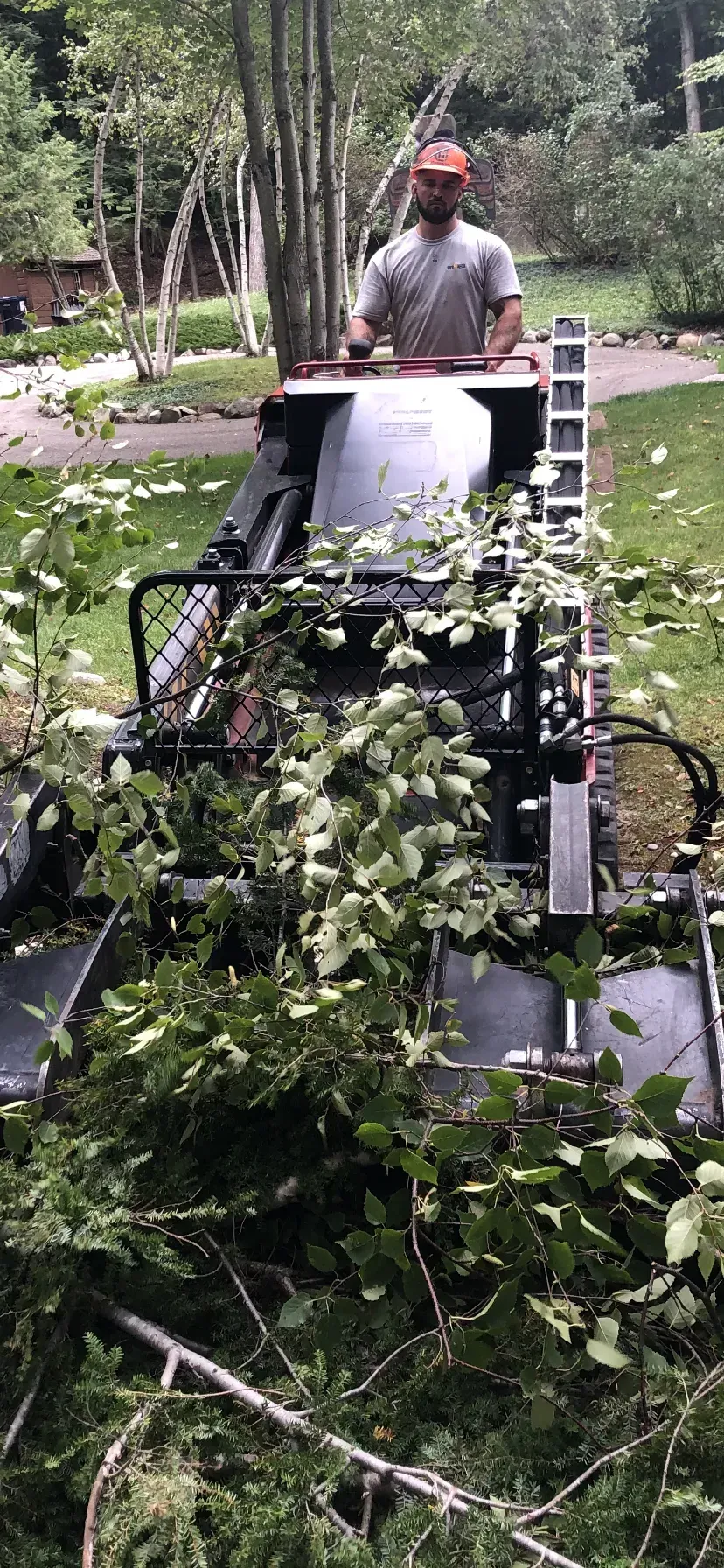 Man in red hat operating a wood chipper. Green foliage being processed. Outdoors setting.