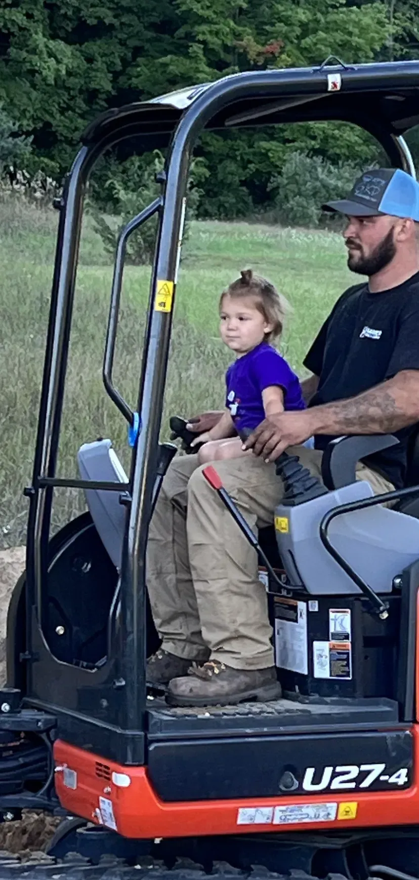 A man and a child in an excavator. The child sits on the man's lap, operating the controls.