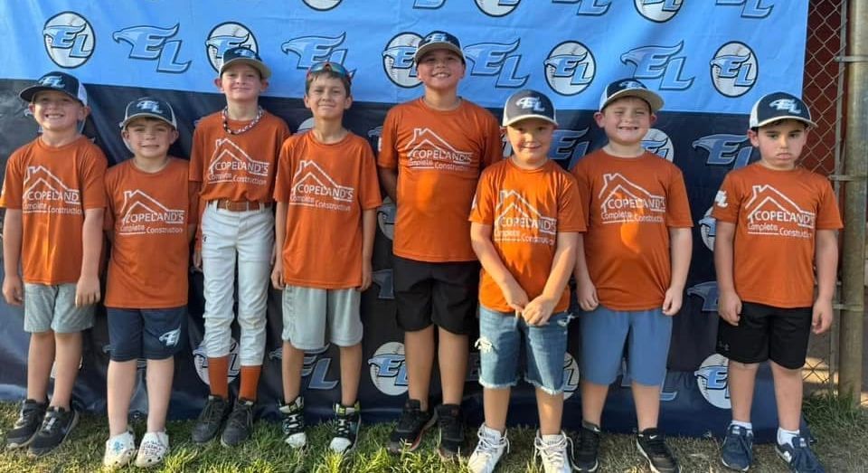 Youth baseball team in orange shirts and hats poses in front of a blue banner.