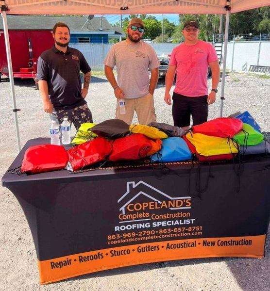 Three men at Copeland's Roofing booth with colorful drawstring bags. Outdoors, overcast.