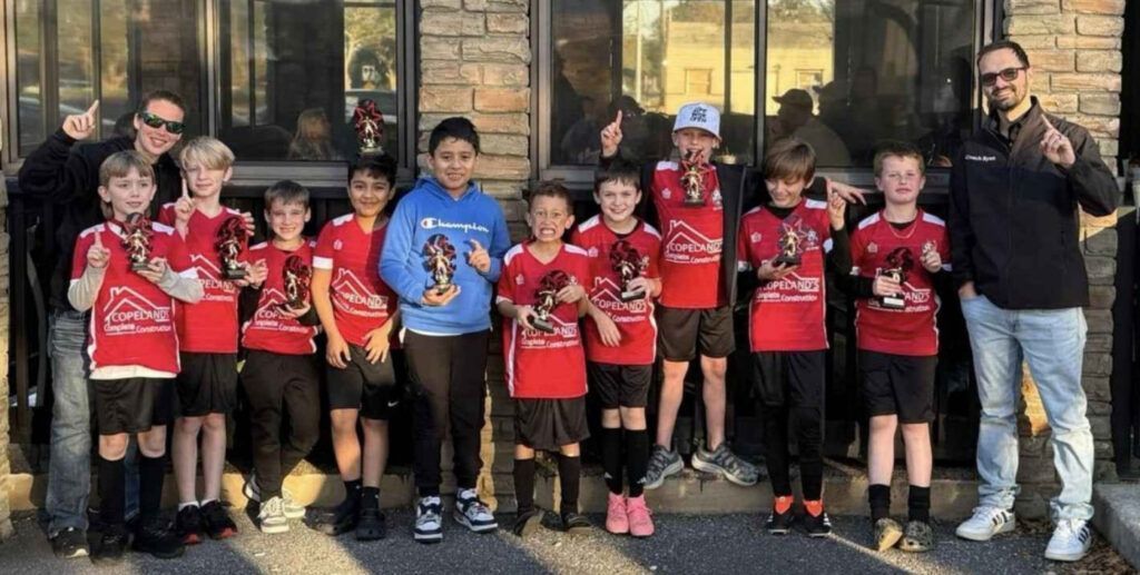 Youth soccer team in red uniforms holding trophies, posing outside a building.