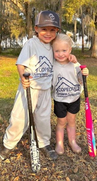 Two young children with bats, wearing matching shirts, smiling and hugging.