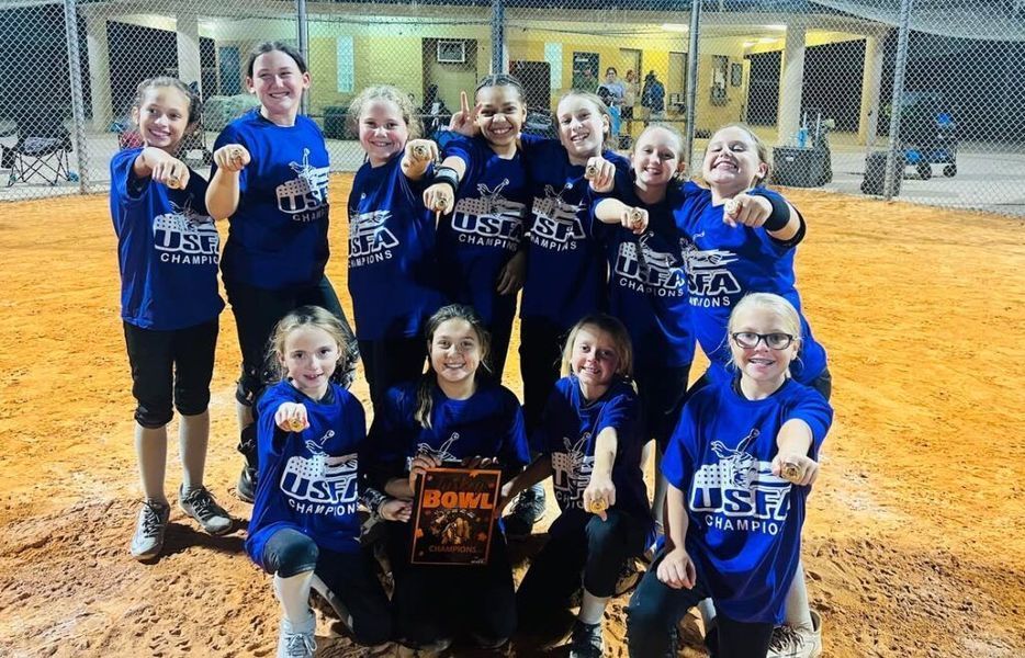 Youth softball team in blue uniforms celebrates with trophy, fists raised on a softball field.