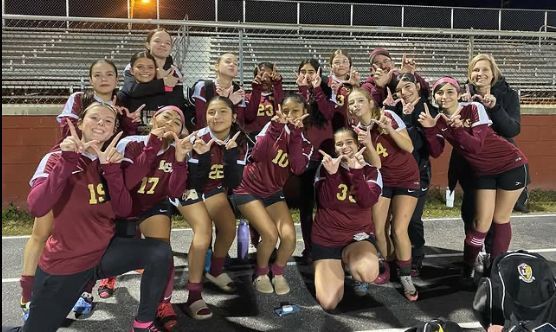 High school girls' field hockey team poses together, making peace signs. They wear maroon and gold uniforms.