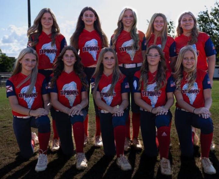 Softball team in red and blue uniforms, kneeling, smiling on a field.