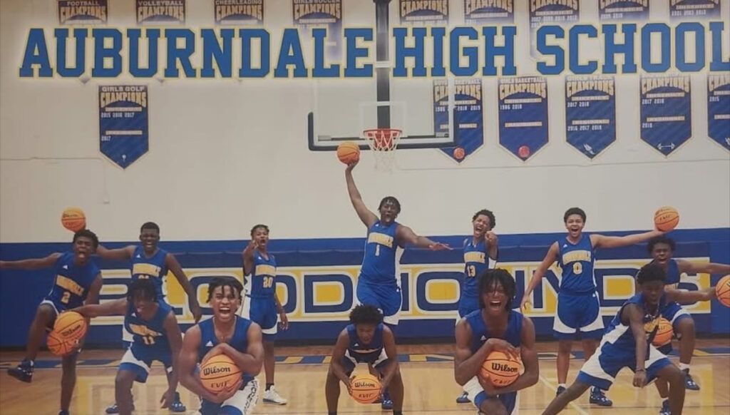 Auburndale High School basketball team poses, holding balls, in blue/gold uniforms; in a gym.