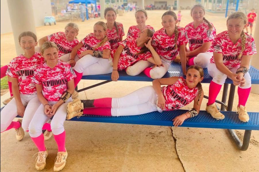 Girls softball team in pink camo shirts and white pants pose on a bench.