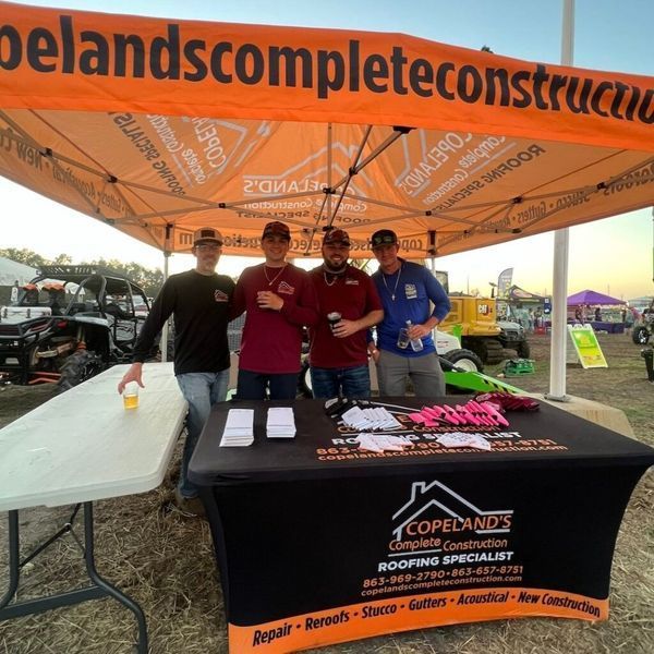 Four men at a construction booth, under an orange tent. Black table, sign, handouts. Outdoor event, daytime.