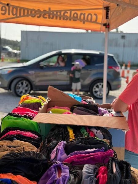 A table with colorful hair accessories; a car with people reaching out; outdoor setting.