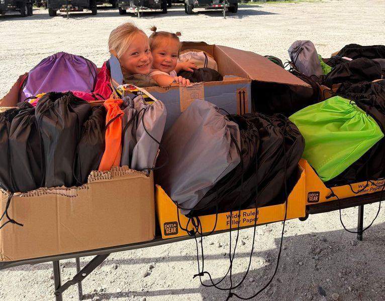 Two young girls smiling, peeking from a cardboard box surrounded by colorful drawstring bags on a table.