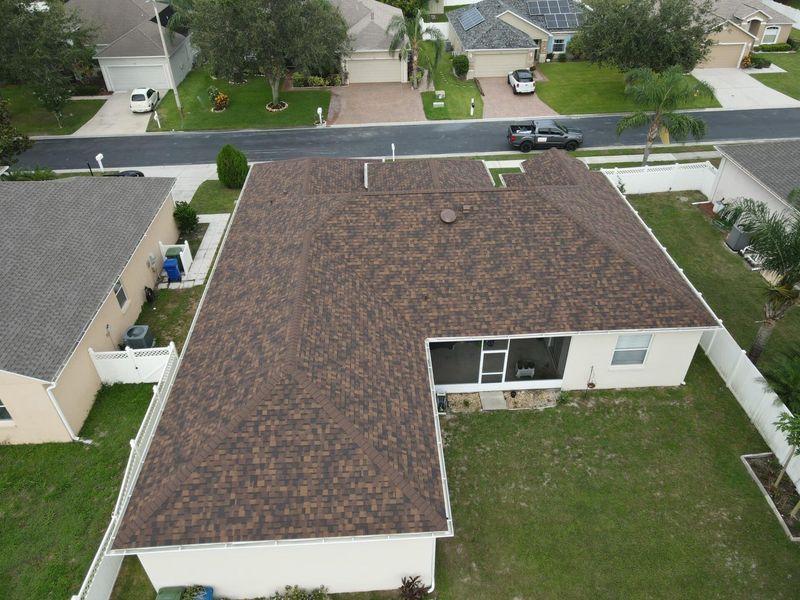 Aerial view of a brown shingle roof on a house in a suburban neighborhood, with a green lawn.