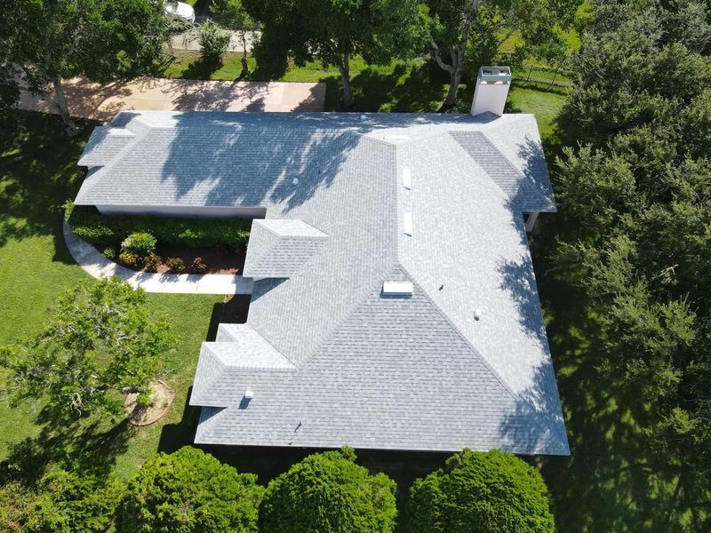 Overhead view of a house with a gray shingle roof, surrounded by green trees and grass.