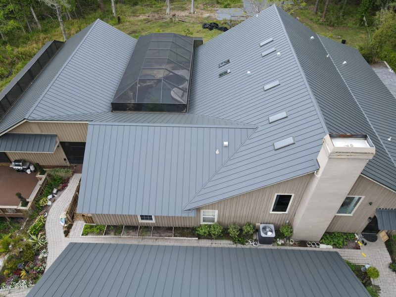 Gray metal roof on a multi-section house with a chimney, skylight, and surrounding landscaping.