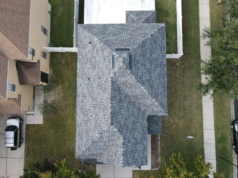 Overhead view of a house with gray shingled roof, surrounded by grass, sidewalk, and other houses.