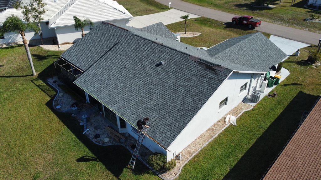 Aerial view of a house with a gray shingle roof and white walls, surrounded by green grass.