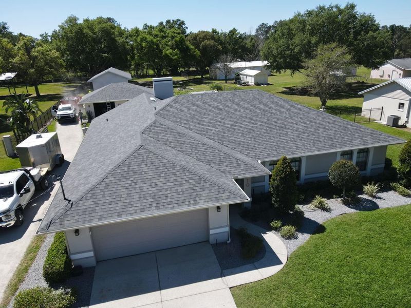 Gray-roofed house with garage, driveway, and surrounding green lawn. White truck parked nearby. Clear blue sky.