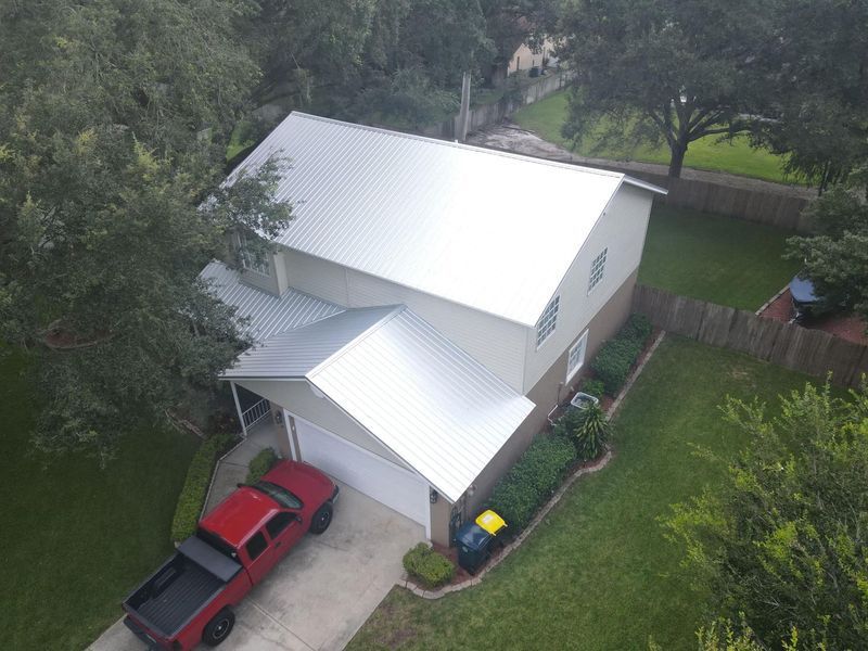 Aerial view of a house with a shiny silver metal roof, red truck in the driveway, green lawn, and trees.