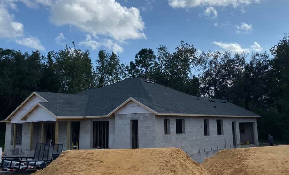 House under construction; gray block walls, new roof, and sand in the foreground, blue sky.