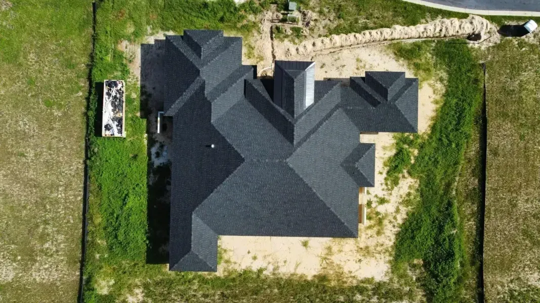 Overhead view of a house under construction with a dark gray roof, surrounded by a grassy, fenced lot.