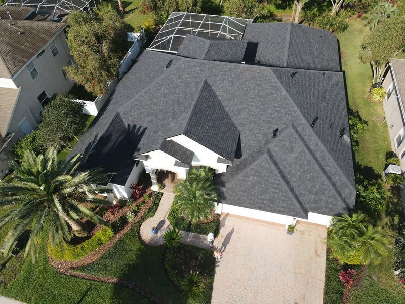 Aerial view of a house with a dark gray roof, light brown driveway, and well-manicured lawn.