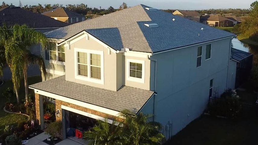 Two-story house with light blue siding, gray roof, and surrounded by palm trees and other houses.