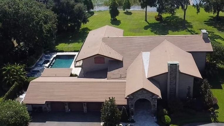 Aerial view of a large beige house with a pool, driveway, and green lawn near a lake.