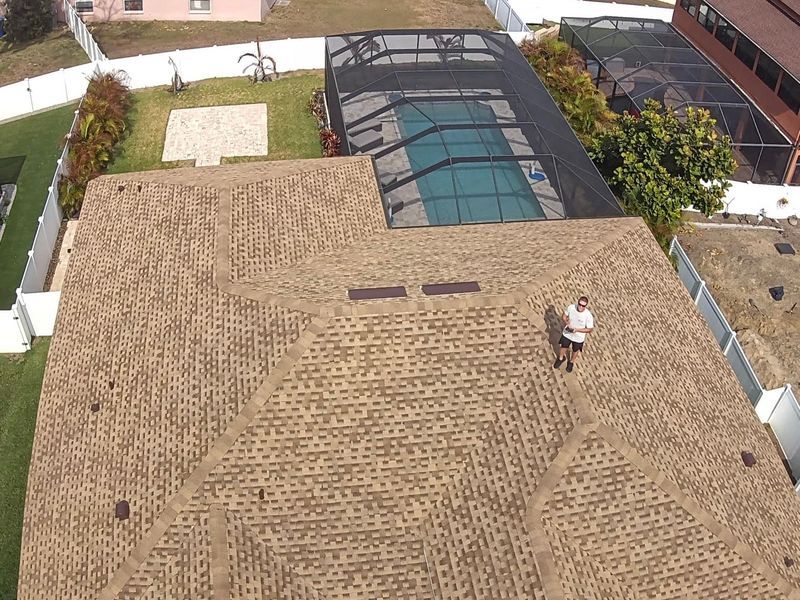 Person standing on a textured brown roof, with a pool and houses in the background.