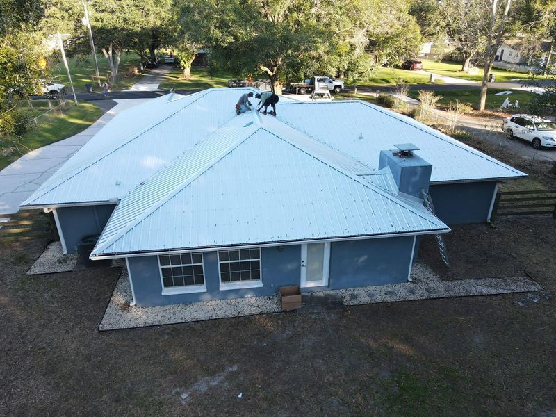 Two roofers install a light blue metal roof on a one-story blue house in a suburban setting.