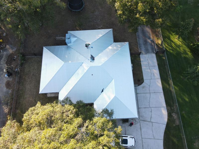 Overhead view of a house with a reflective metal roof and a long concrete driveway, surrounded by trees.