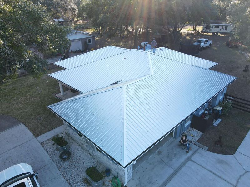 Aerial view of a house with a shiny metal roof under sunlight; a vehicle and trailer are visible in the background.