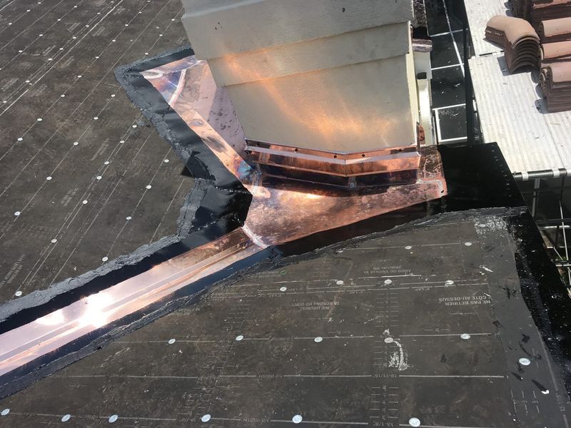 Copper flashing installed around a chimney on a dark asphalt roof, with a copper gutter.