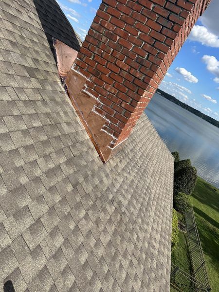 Rooftop with brown shingles, brick chimney, and lake view under a partly cloudy sky.