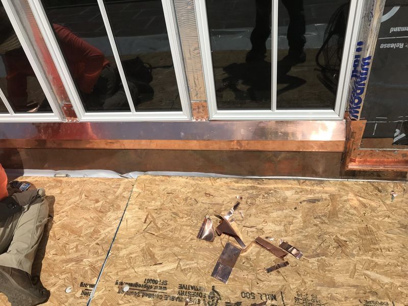 Copper flashing being installed around a window frame on an unfinished plywood surface.