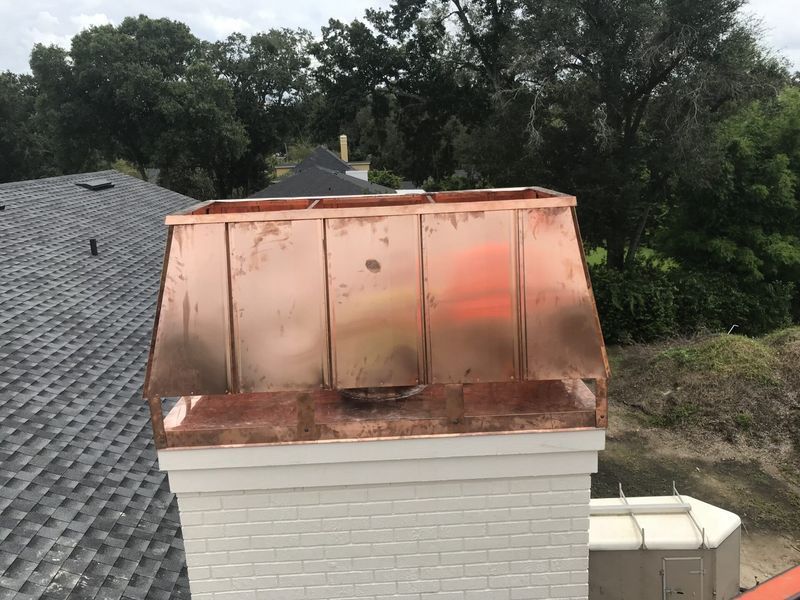 Copper chimney cap on a white brick chimney, with dark gray shingles on the roof.