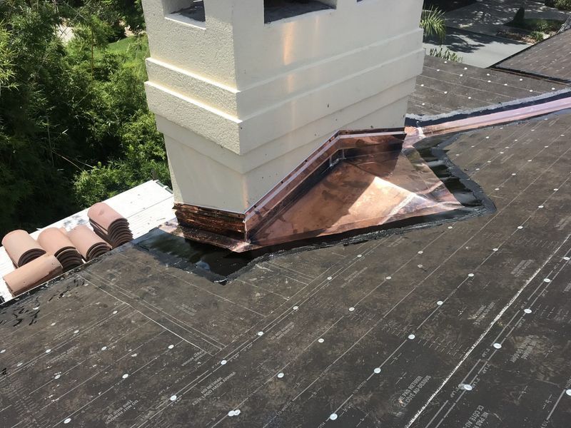 Chimney on a dark roof with copper flashing, surrounded by trees.