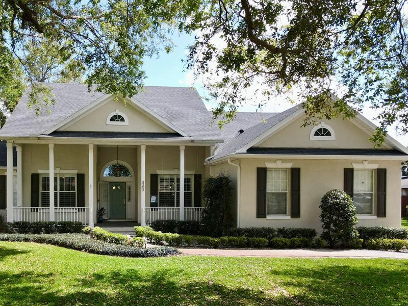 Beige house with dark shutters, white columns, and green lawn.