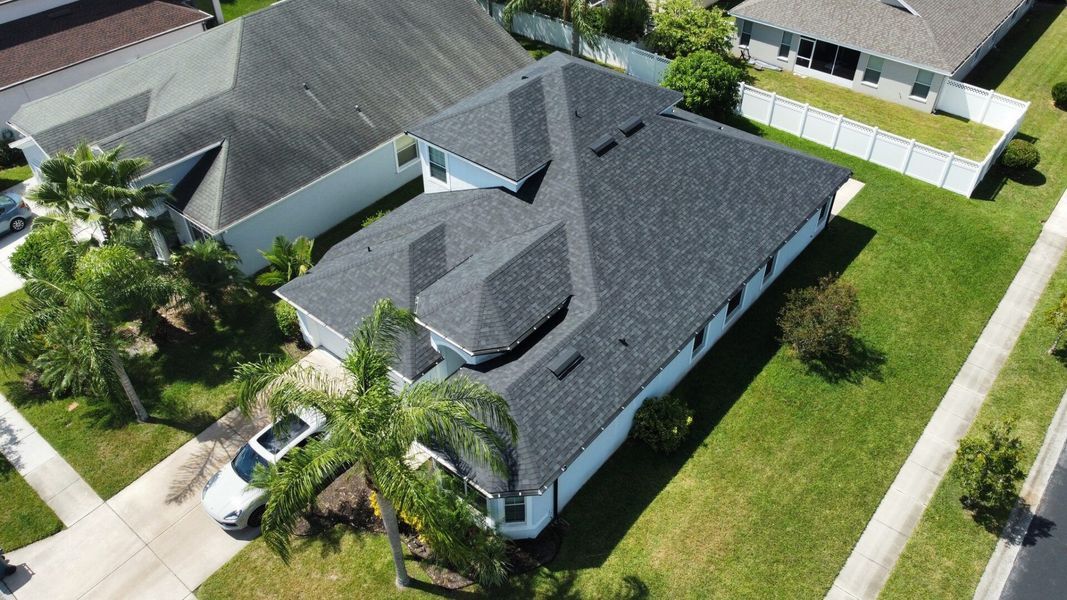 Aerial view of a house with a dark gray roof, surrounded by green lawn and palm trees.
